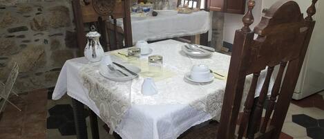 Una mesa de comedor con mantel blanco y cubertería, rodeada de paredes de piedra y una silla de madera con cojín de asiento tejido.