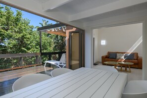 Et moderne udendørs opholdsområde med træterrasse, hvide møbler og en glasdør, der fører til en balkon med udsigt over træer.