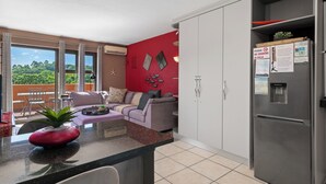 Cozinha moderna com geladeira, sala de jantar com mesa e cadeiras, e sala de estar com sofá e planta.