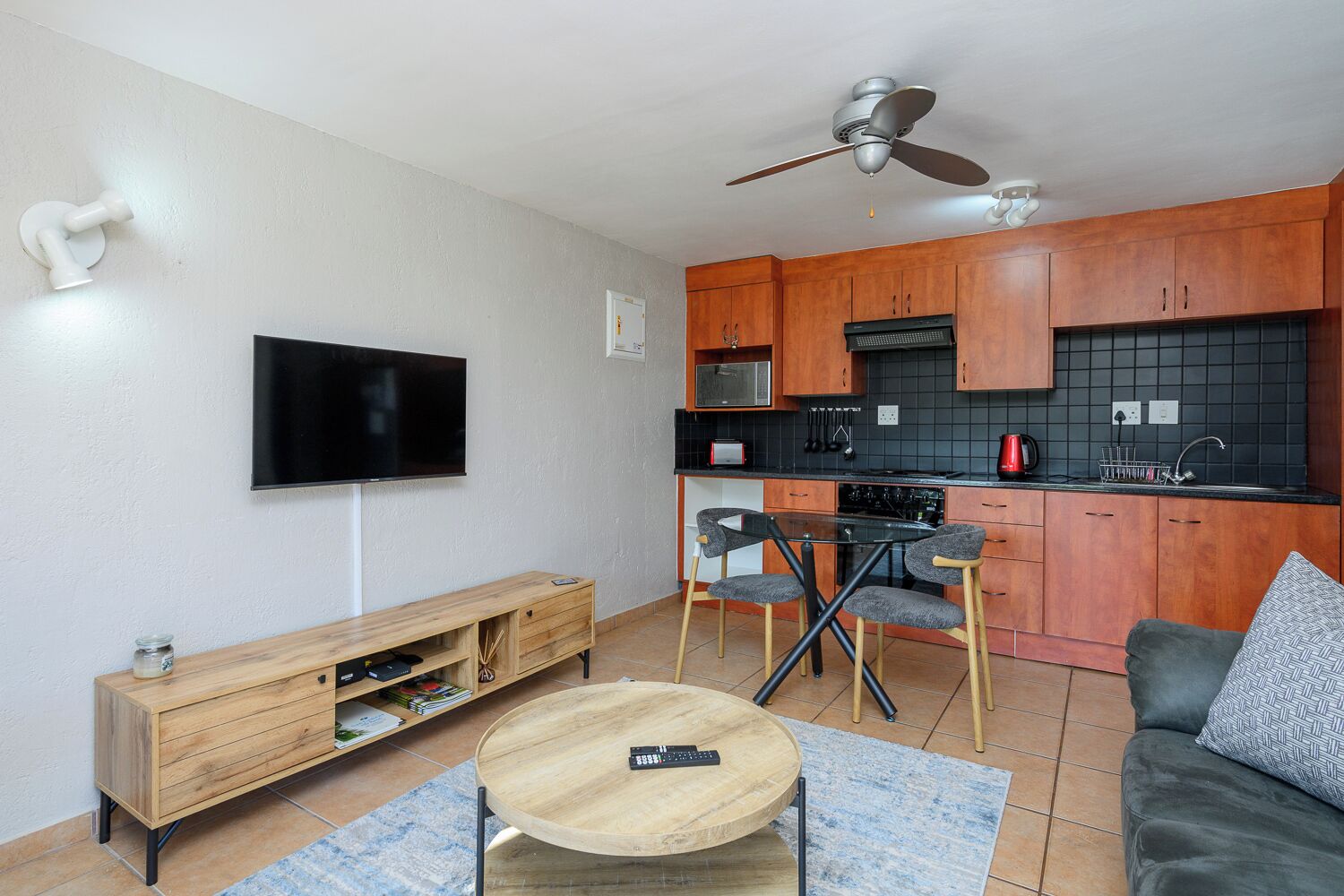 A modern living room with a flat-screen TV, wooden furniture, a dining area with chairs, a sofa, and a ceiling fan.