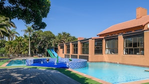 A swimming pool with a slide, surrounded by palm trees and a building with large windows. - San Lameer Villa Rentals One Bedroom Standard 10425 (Southbroom)