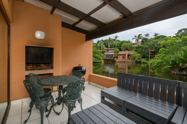 An outdoor patio with a table and chairs, a barbecue grill, and a view of a lake and surrounding greenery.