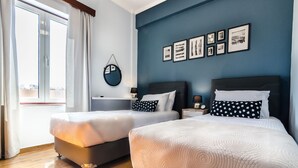 A hotel room with two beds, a large window with curtains, a wall with framed pictures, and a round mirror. - Casa Ithomi (Chania)