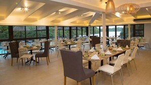 Un vaste espace dédié à la salle à manger, avec de nombreuses tables et chaises, de grandes fenêtres offrant une vue sur l’extérieur et un plafond haut avec des poutres apparentes.