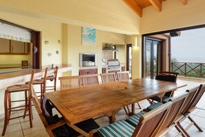 Un espace repas avec une grande table et des chaises en bois, une cuisine équipée d’un barbecue et une vue sur l’extérieur.