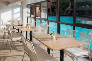 A poolside dining area with tables and wicker chairs, overlooking a swimming pool.