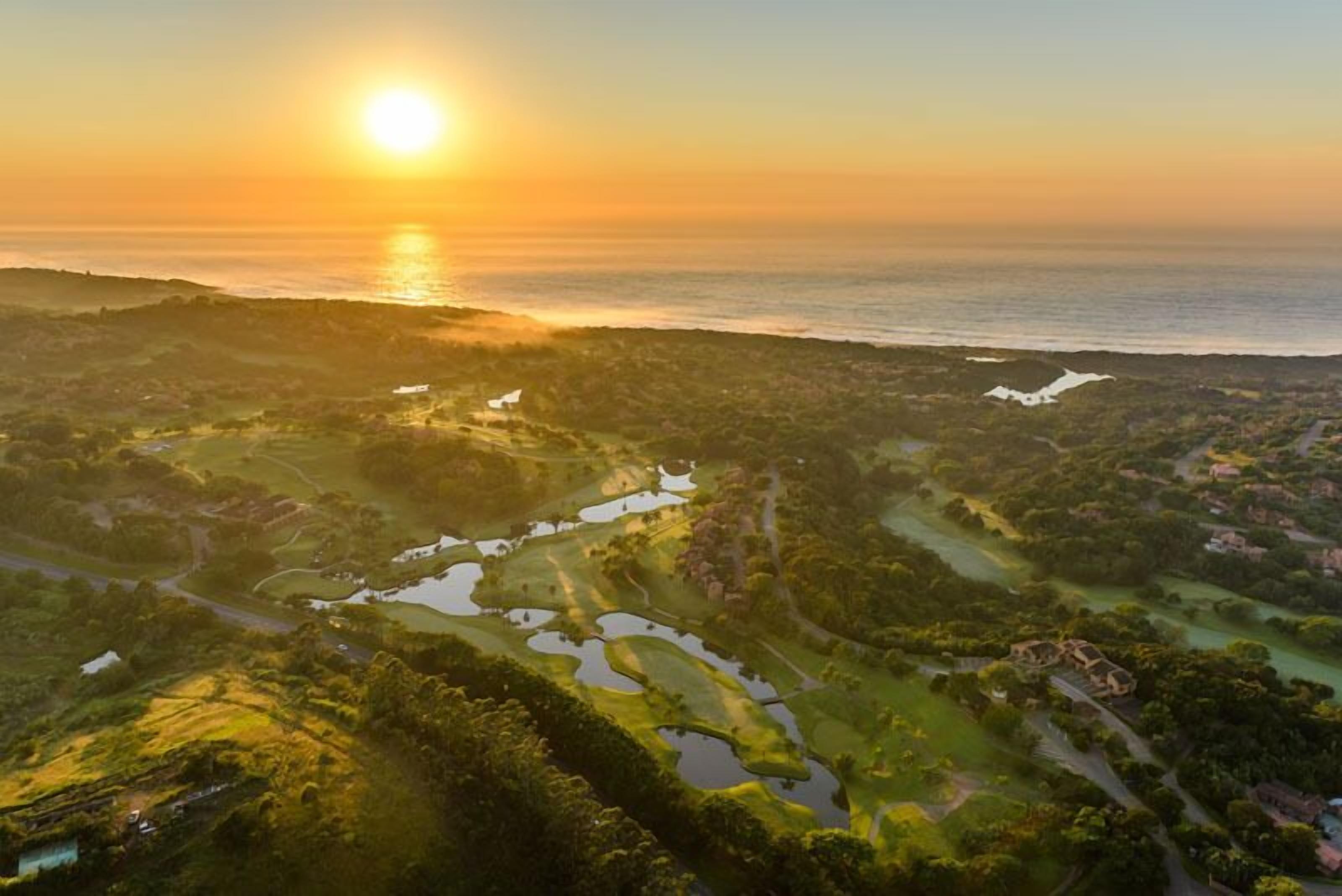 An aerial view of a golf course with water hazards and a sunset in the background.