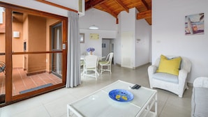 Ein geräumiger Wohnbereich mit einer Schiebeglastür, die auf einen Balkon führt, ein weißes Rattangeflechtsofa mit einem gelben Kissen, ein Glastisch und ein Essbereich mit weißen Stühlen und einem Tisch.