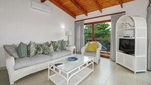 Ein Wohnzimmer mit einem weißen Rattan-Sofa, einem Glastisch, einer weißen Unterhaltungseinheit und einem großen Fenster mit Vorhängen.