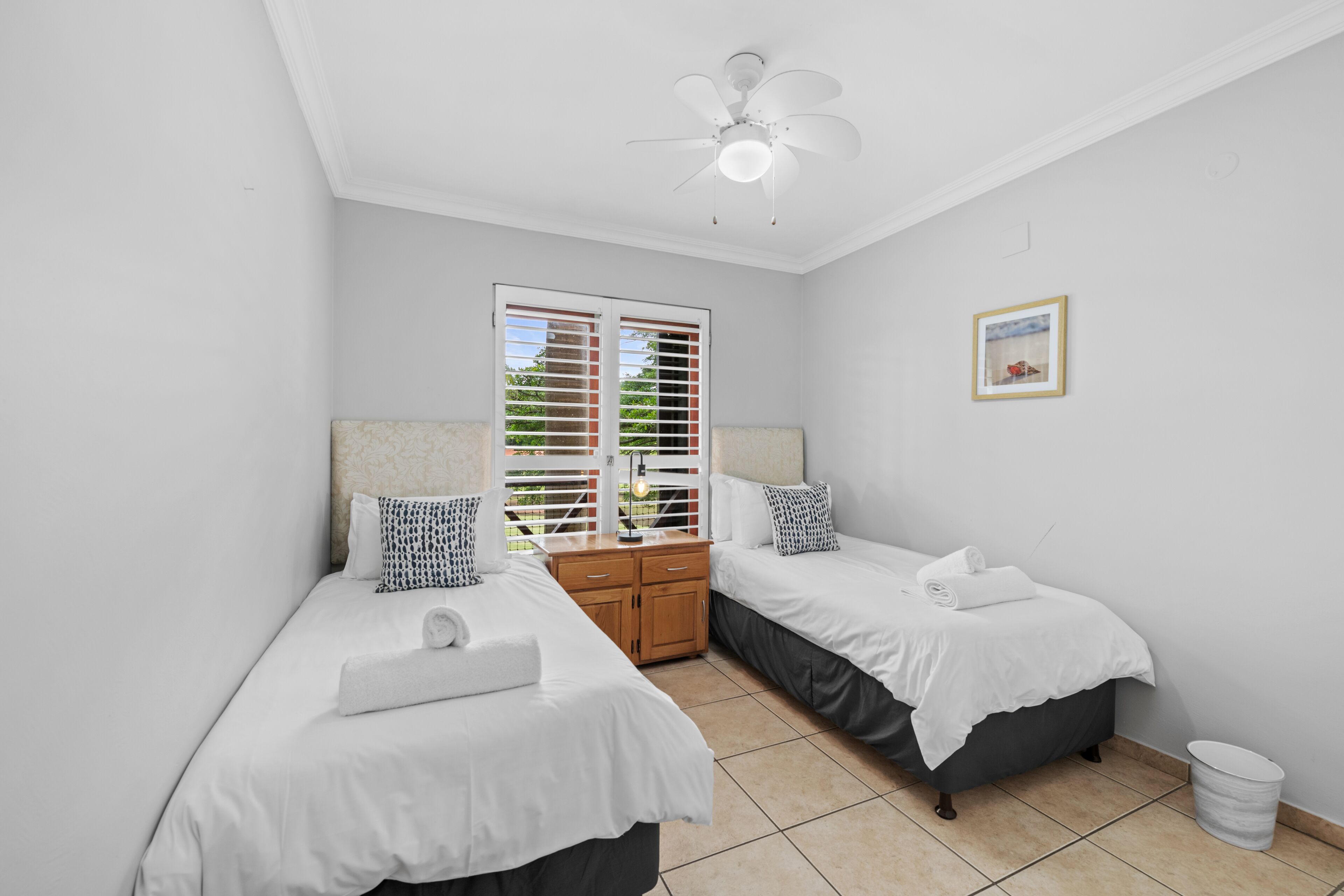A bedroom with two beds, a ceiling fan, a window with blinds, a bedside table, and a picture on the wall.