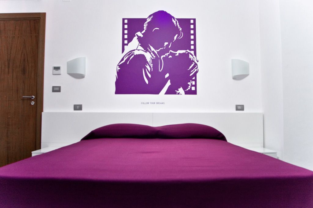 Et soveværelse med lilla sengetæppe, en vægmonteret lampe og et billede med teksten "FOLLOW YOUR DREAMS".