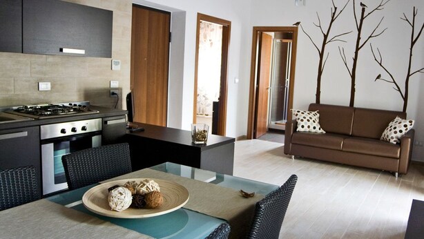 A modern kitchen with a gas stove, dining table with chairs, and a sofa in a living area with a view of the outdoors. - InStile aparthotel (Ascea)