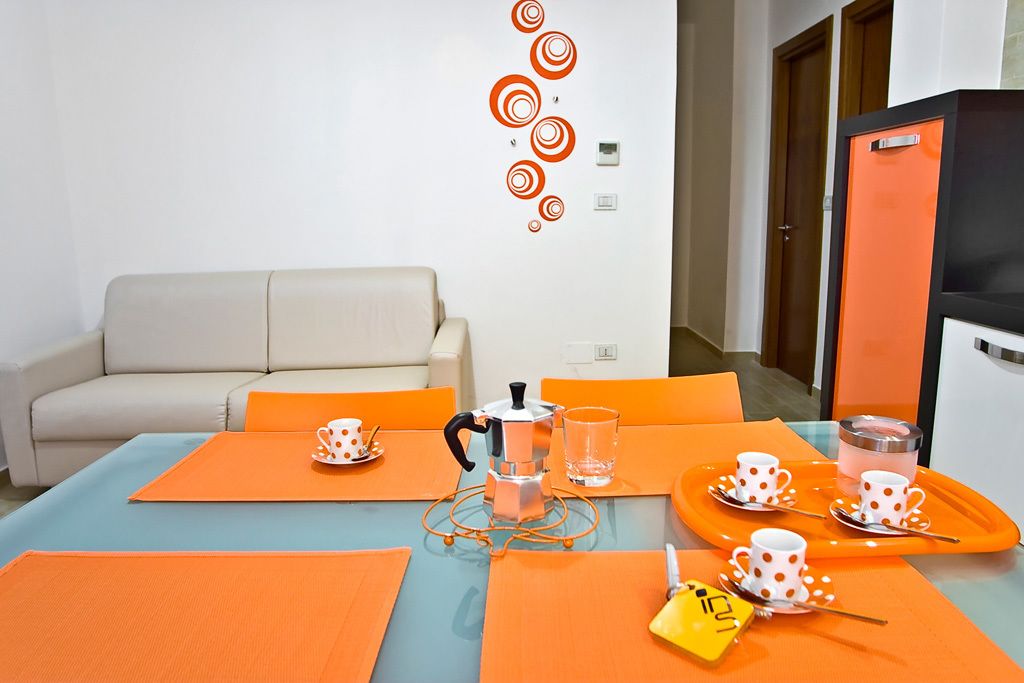 A modern hotel room with a beige sofa, orange and blue accent pillows, a coffee table with a coffee pot and cups, and a bright orange cabinet.