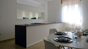 A modern kitchen with a central island, a dining table with chairs, and a window with sheer curtains. - InStile aparthotel (Ascea)