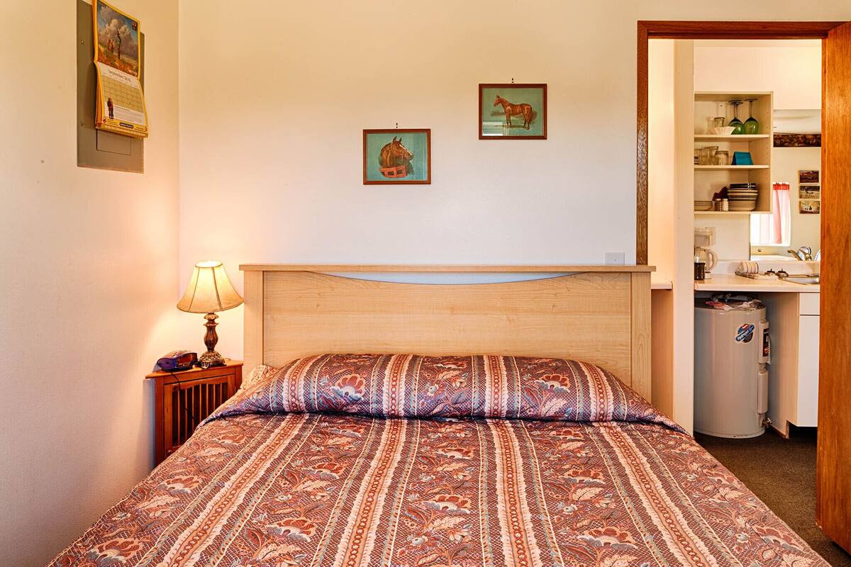 Quarto com cabeceira de madeira, colcha estampada, mesa de cabeceira com abajur e duas fotos emolduradas na parede.