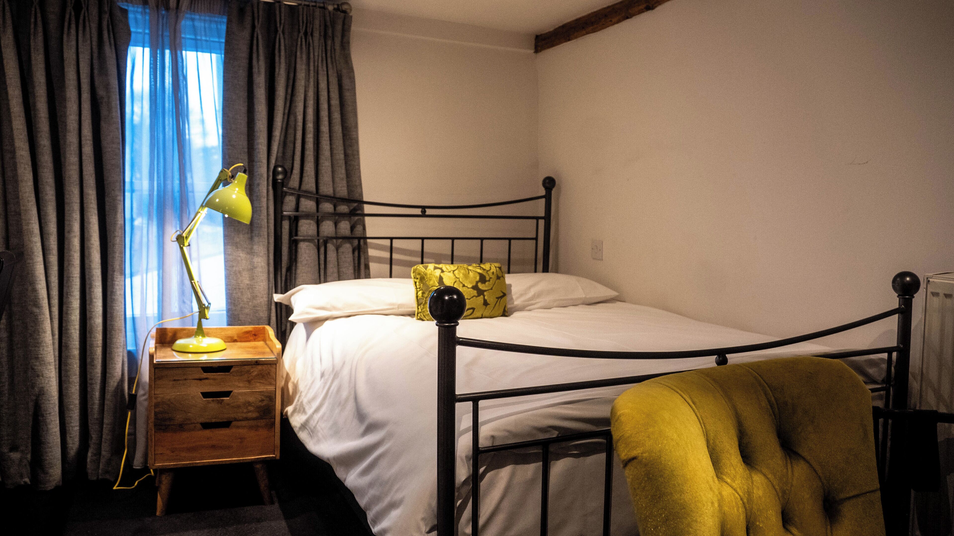 Un lit simple avec un cadre en métal, une table de chevet avec une lampe, une chaise et une fenêtre avec des rideaux.