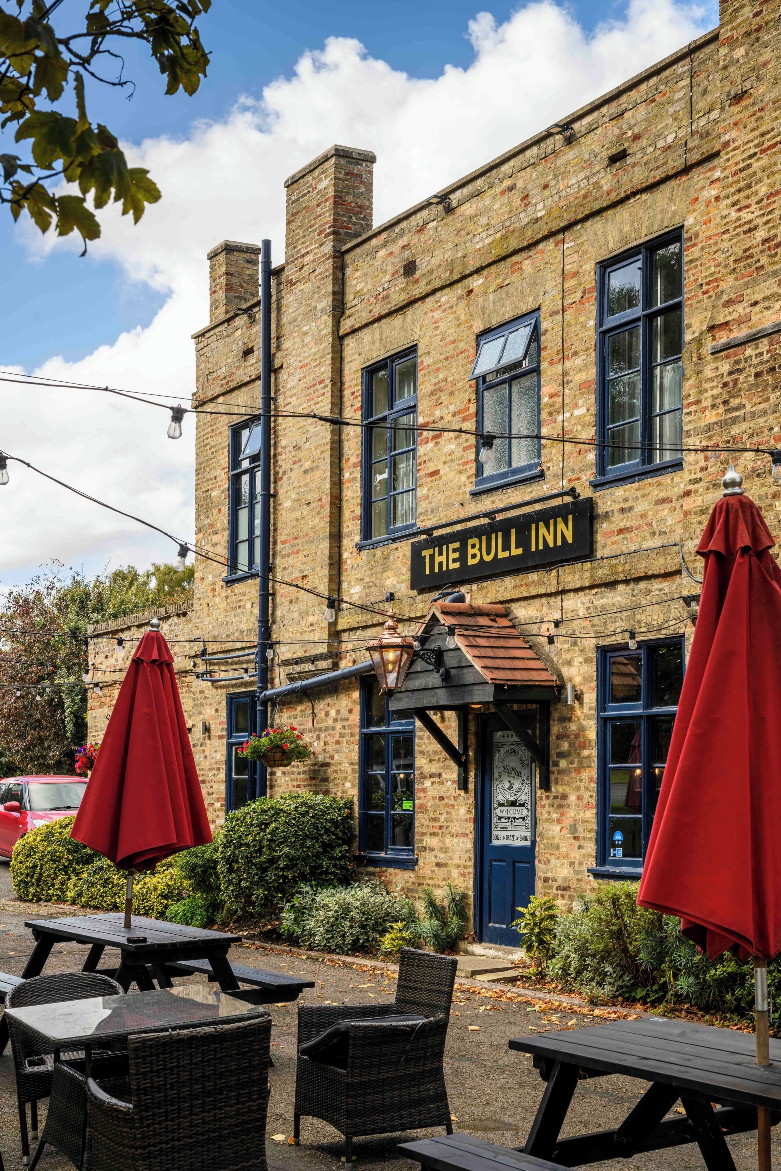 Un edificio de ladrillo con un letrero que dice 'The Bull Inn'. Hay mesas y sillas al aire libre, y un paraguas rojo.