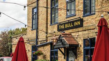 Un edificio de ladrillo con un letrero que dice 'The Bull Inn'. Hay mesas y sillas al aire libre, y un paraguas rojo.