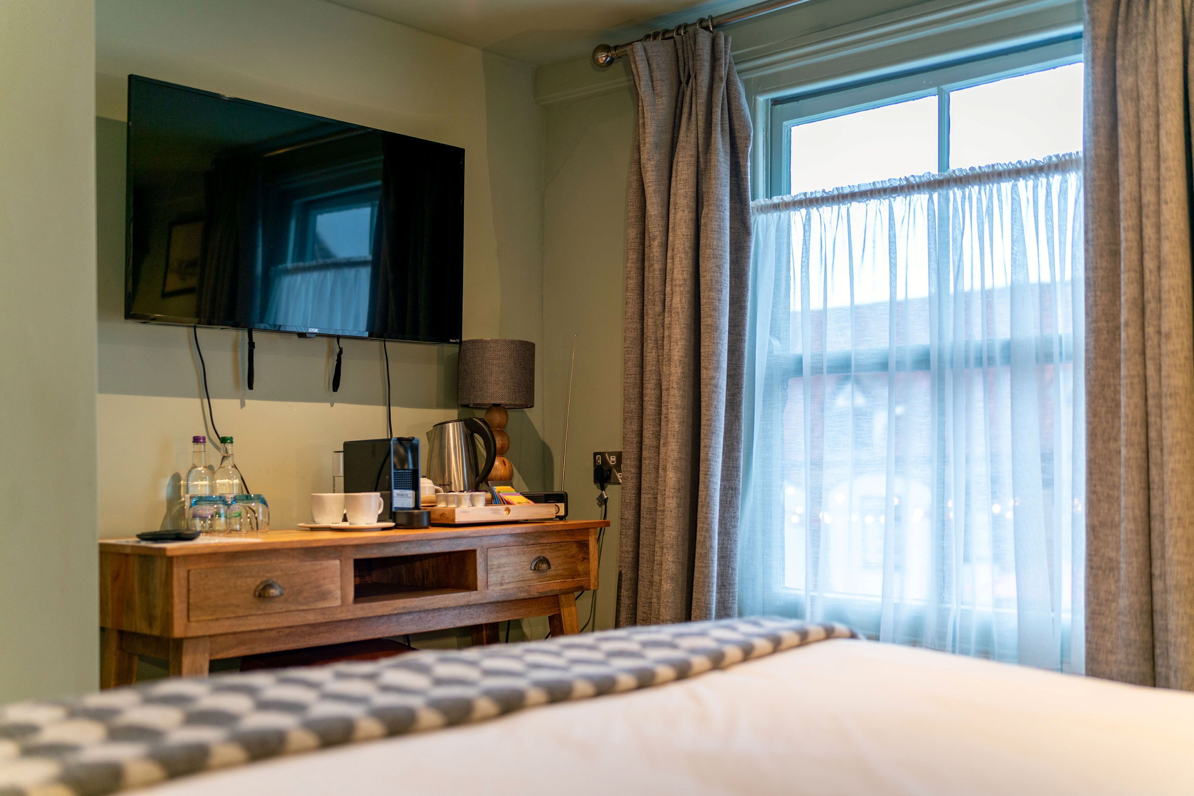 A hotel room with a bed, a wooden nightstand, a flat-screen TV mounted on the wall, a window with curtains, and a view of the outside.