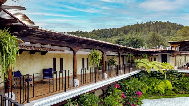 A covered outdoor area with wooden benches and a railing, surrounded by lush greenery and a view of a forested hill. - Casa Faustina Boutique Hotel (Antigua Guatemala)