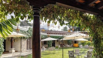 Un patio al aire libre con mesas y sillas, cubierto por una pérgola, rodeado de abundante vegetación.
