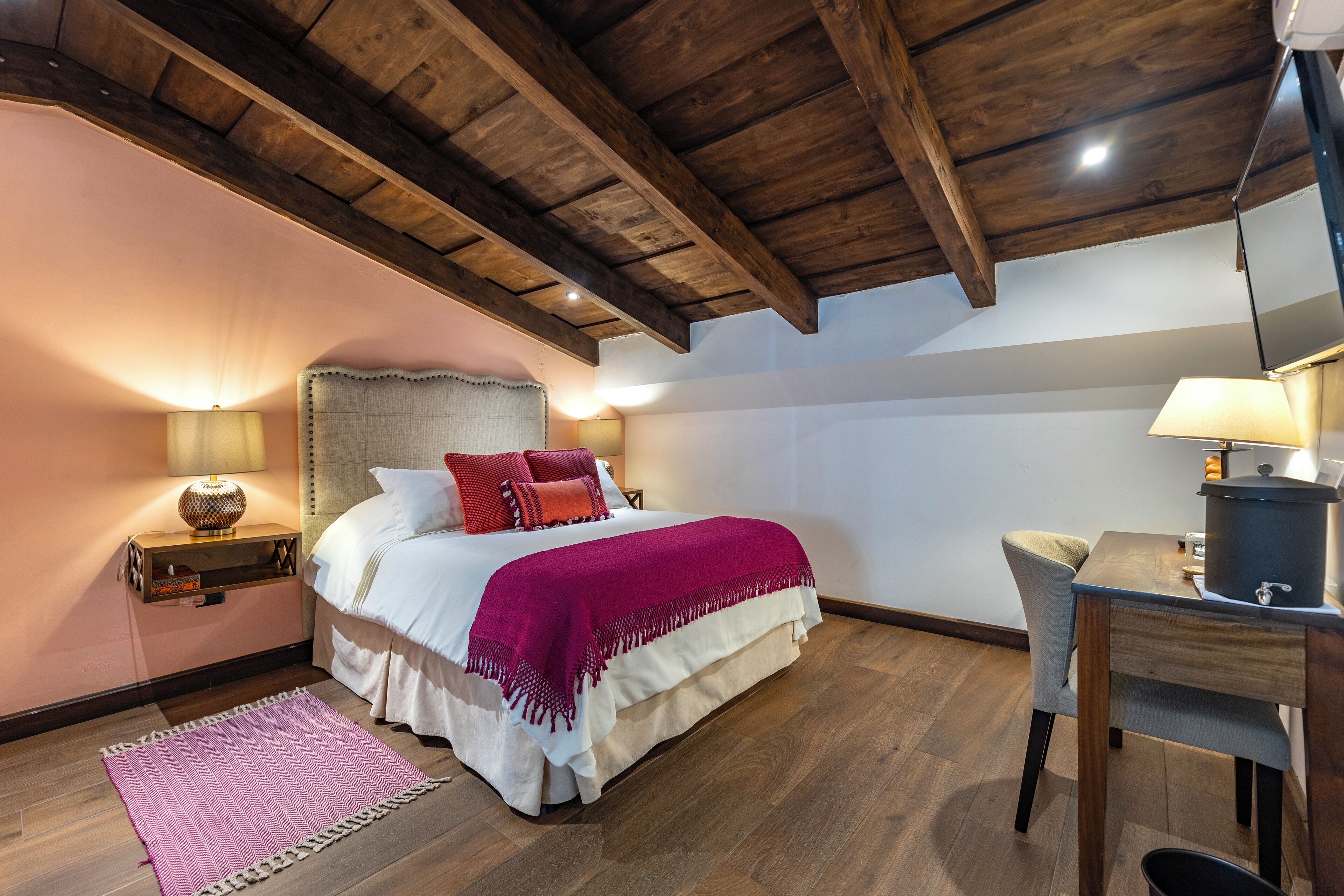 A bedroom with a wooden ceiling, a bed with a white and red bedspread, a nightstand with a lamp, and a desk with a lamp.