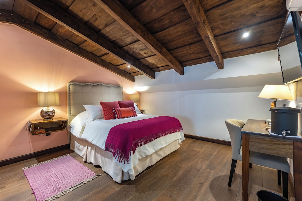 A bedroom with a wooden ceiling, a bed with a white and red bedspread, a nightstand with a lamp, and a desk with a lamp.