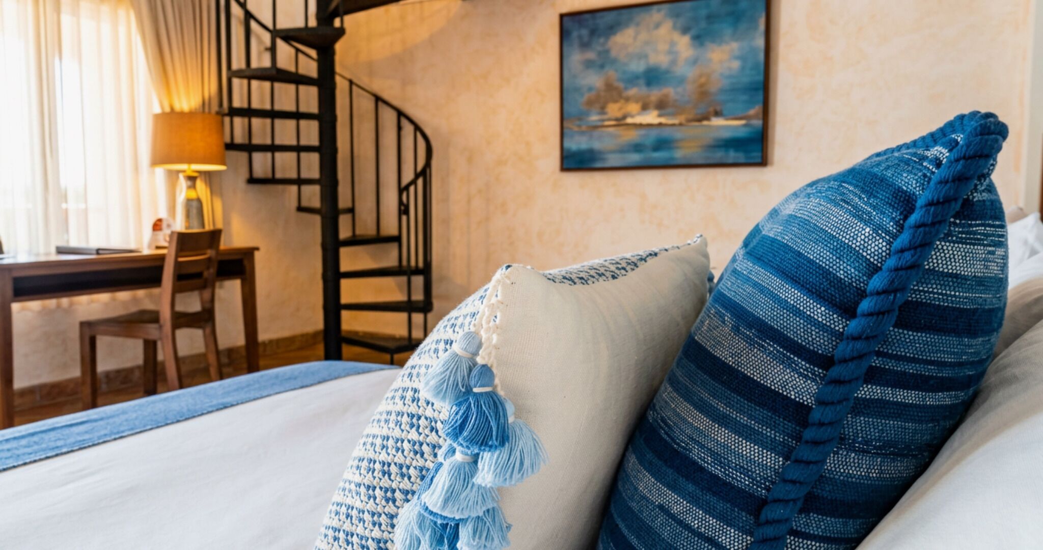 A hotel room with a bed, a desk, a chair, a spiral staircase, and a painting on the wall.