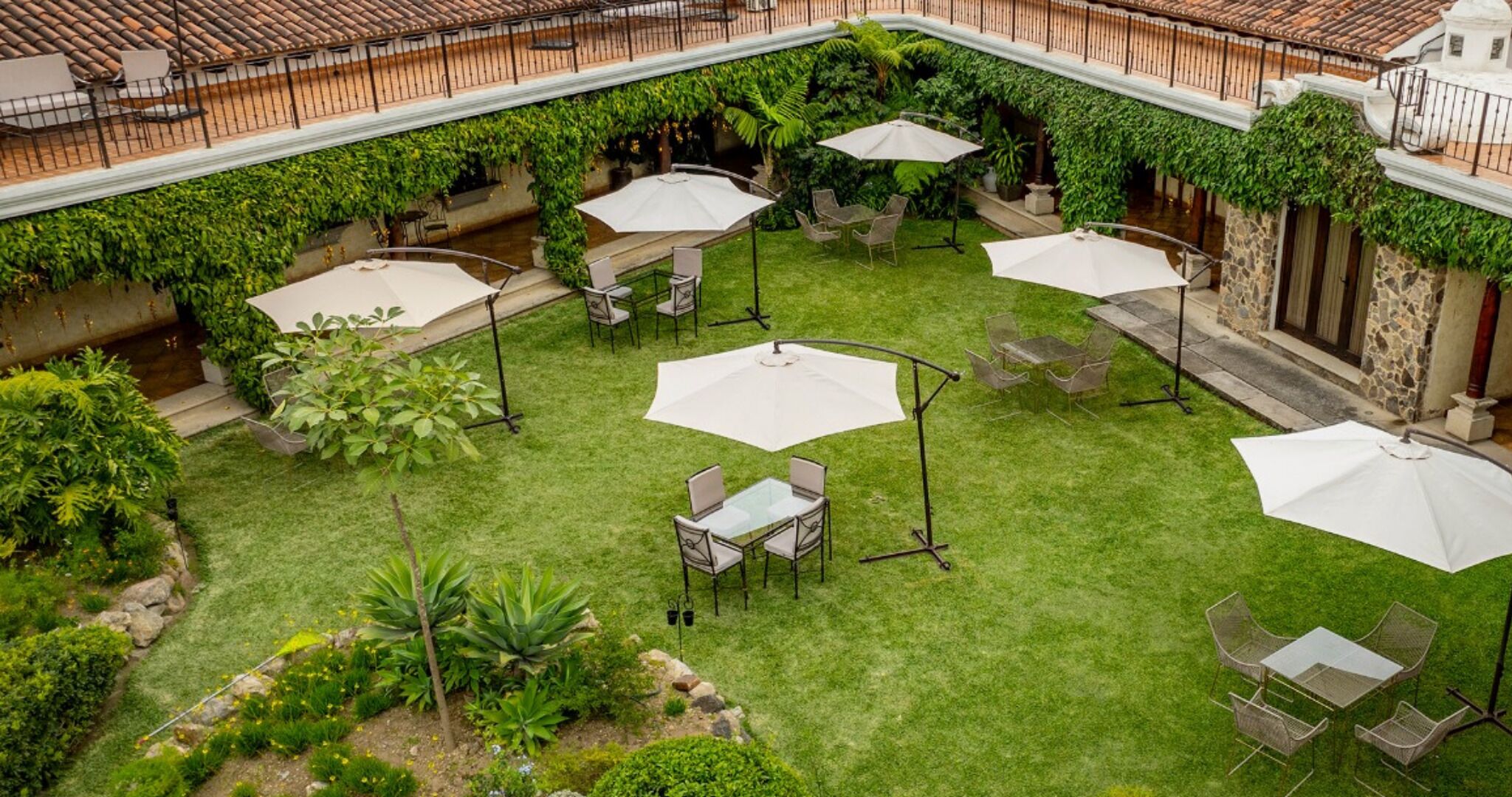 An outdoor patio area with multiple tables and umbrellas, surrounded by lush greenery and stone walls.