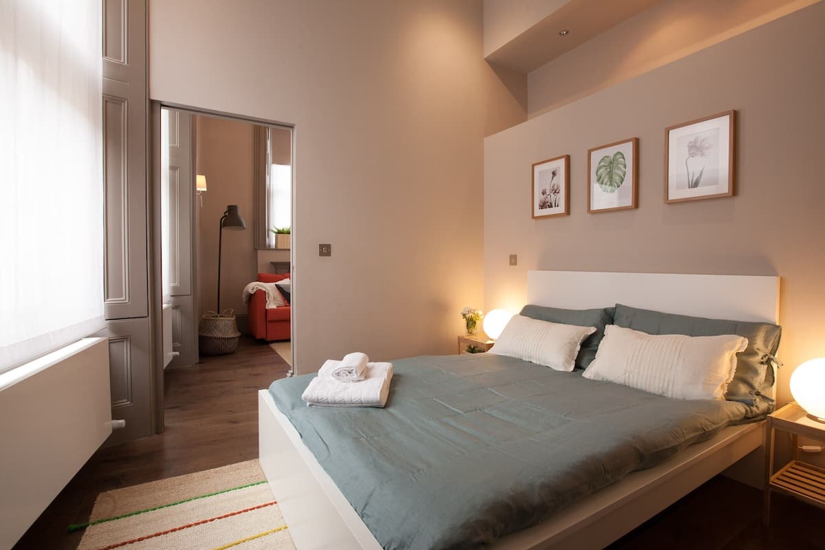 A modern bedroom with a large bed, a bedside table with a lamp, and framed botanical prints on the wall.