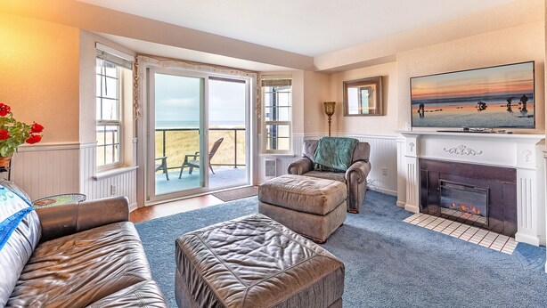 A living room with a fireplace, a sofa, a chair, and a large window with a view of the beach. - Sweet Caroline Inn (Ocean Shores)