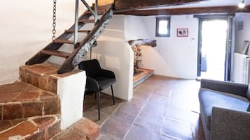 Una escalera de piedra con escalones que conducen a un rellano, una silla negra y un sofá gris en una habitación con suelo de baldosas y vigas de madera.