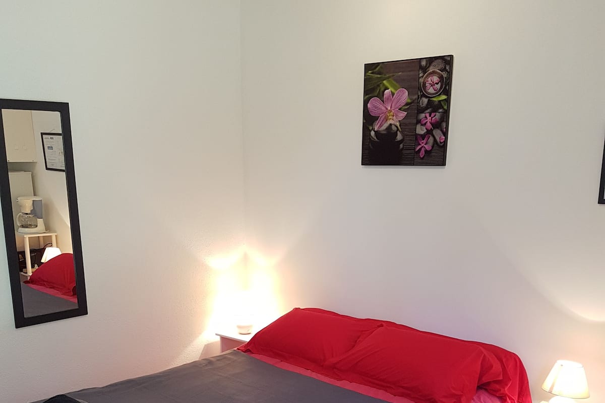 Un lit bien fait, avec une literie rose et grise, une lampe de chevet et une petite table sur laquelle se trouve une autre lampe.