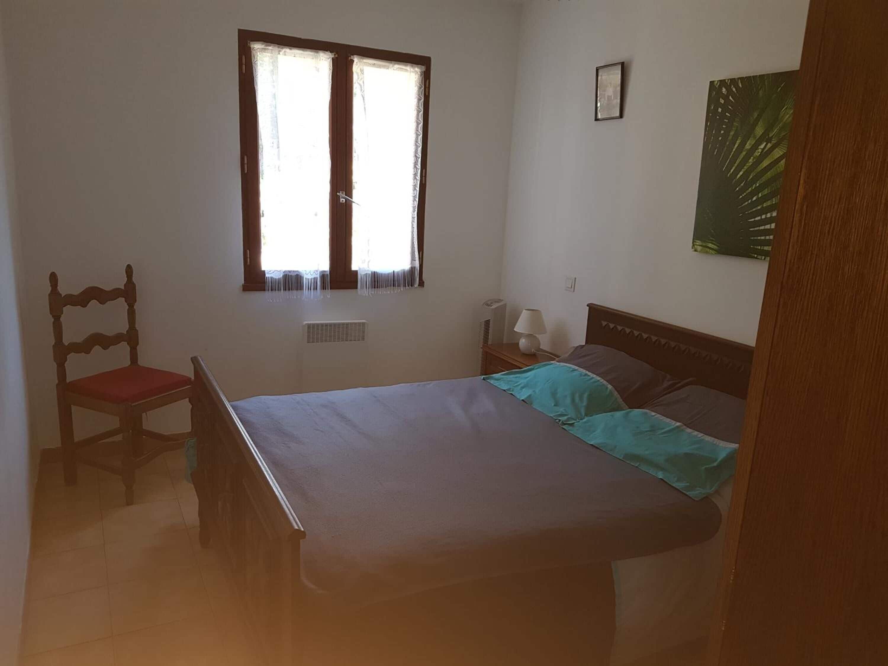 A bedroom with a bed, a chair, a window, a bedside table with a lamp, and a framed picture on the wall.