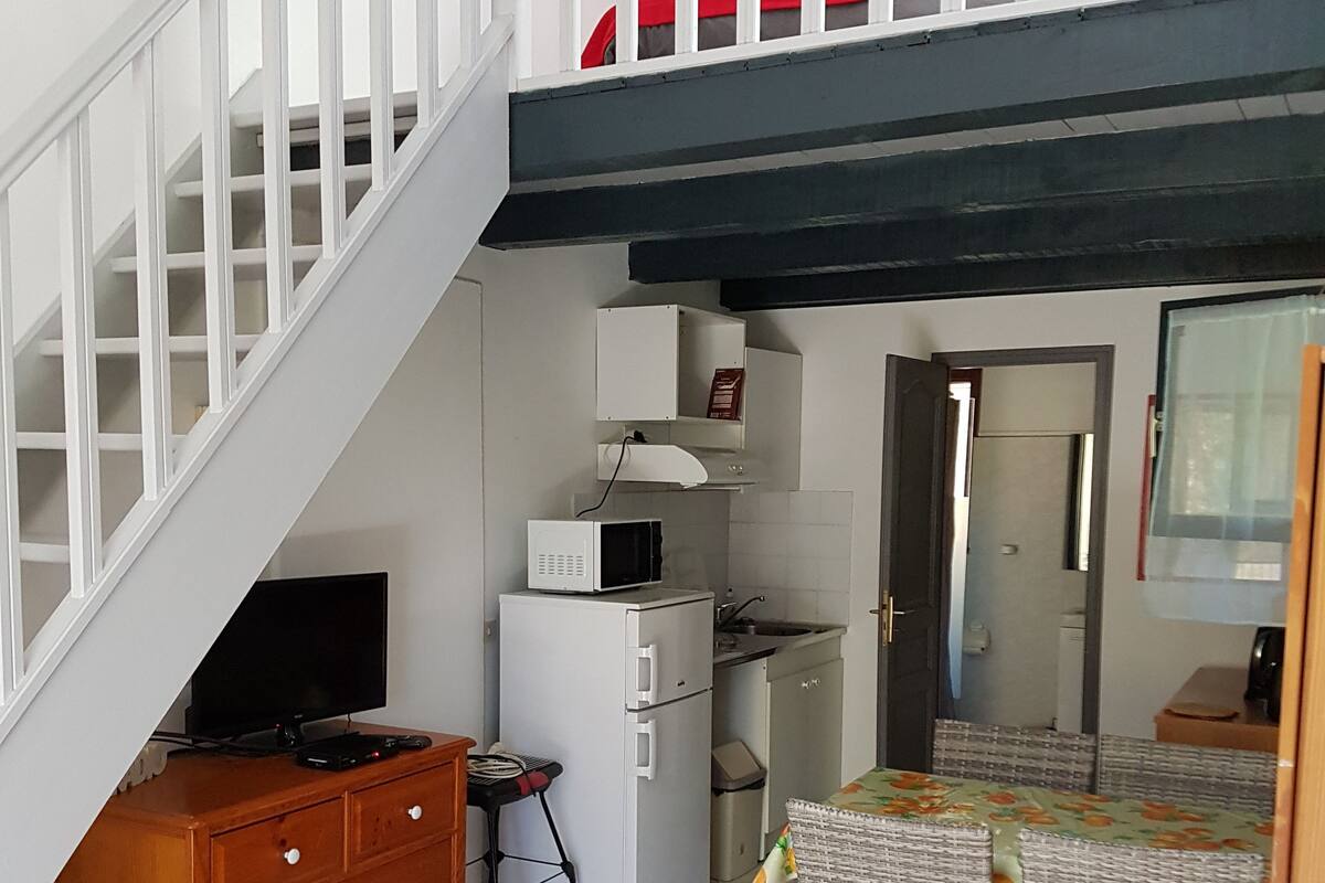 Un espace de vie compact comprenant un escalier, un coin cuisine, un espace repas et un téléviseur posé sur une commode en bois.
