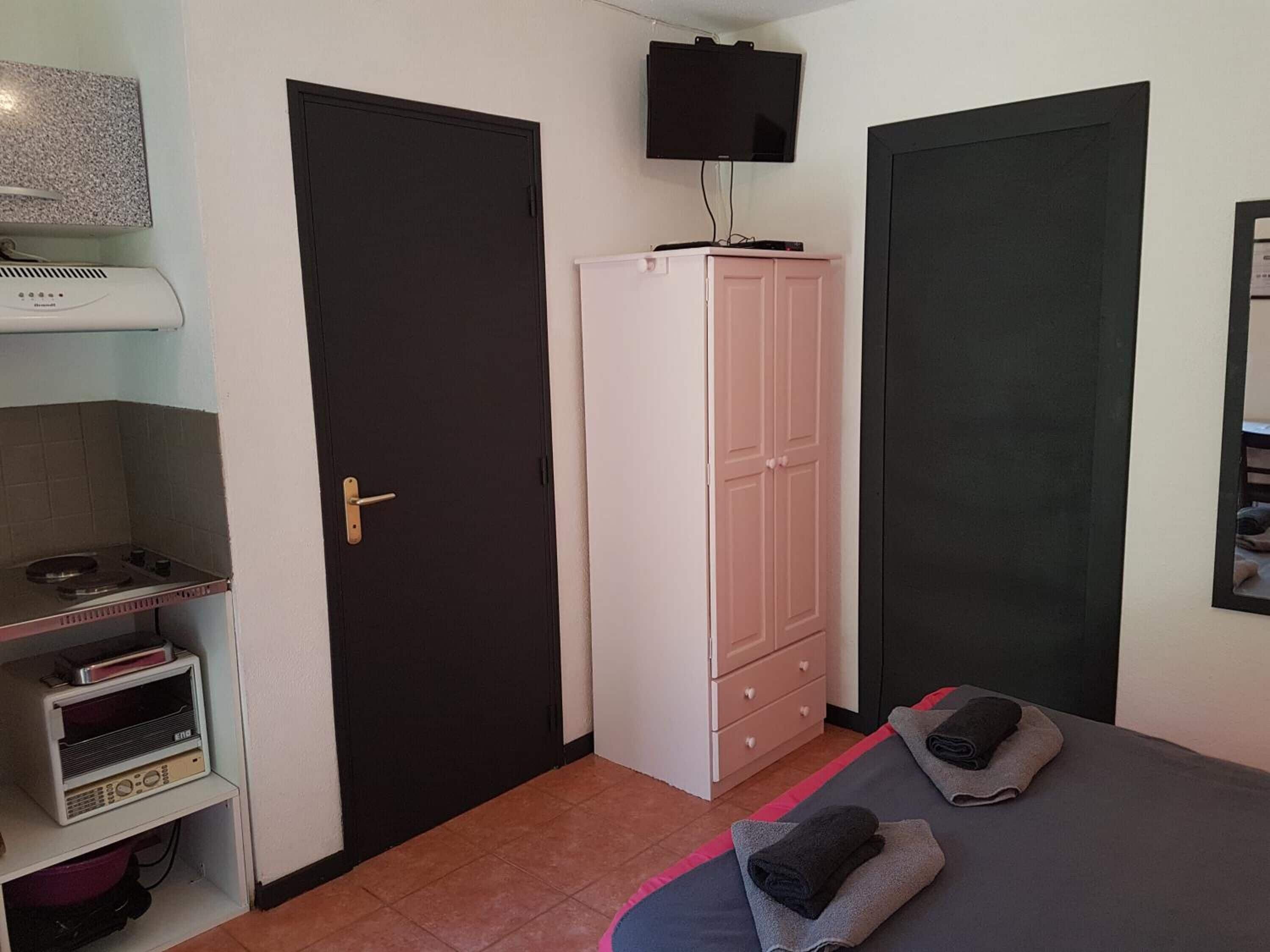 A small, compact room with a bed, a wardrobe, a television, and a kitchenette area.