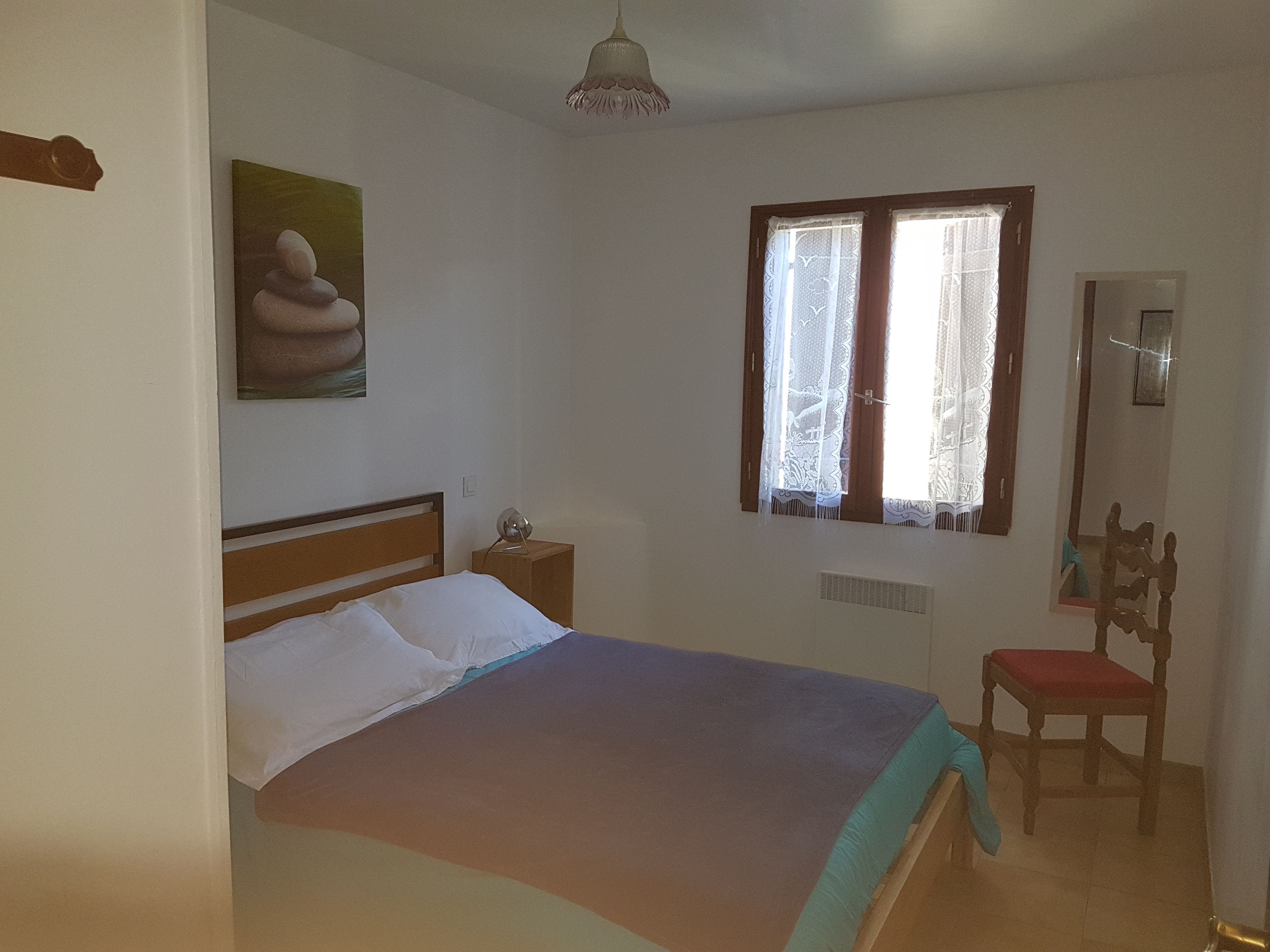 Une chambre à coucher comprenant un lit, une table de chevet, une lampe, une fenêtre avec des rideaux, une chaise et un tableau accroché au mur.