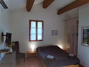 Une chambre à coucher avec un lit, une table de chevet avec une lampe, un bureau avec un ordinateur et un vase de fleurs, une fenêtre avec des rideaux et un tableau encadré au mur.