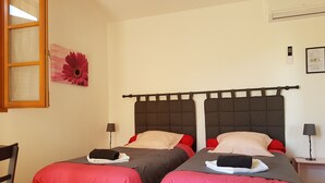 Quarto com duas camas, cada uma com cabeceira escura e roupa de cama vermelha. Há uma janela com moldura de madeira, um ar-condicionado fixado na parede e uma pequena mesa com abajur.