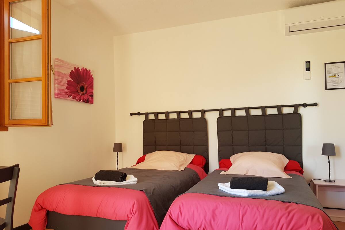 Une chambre avec deux lits, chacun doté d’une tête de lit sombre et d’une literie rouge. On y trouve une fenêtre avec un cadre en bois, un climatiseur fixé au mur et une petite table avec une lampe.