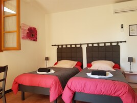 Habitación con dos camas, cada una con cabecera oscura y ropa de cama roja. Se observa una ventana con marco de madera, un aire acondicionado instalado en la pared y una pequeña mesa con lámpara.