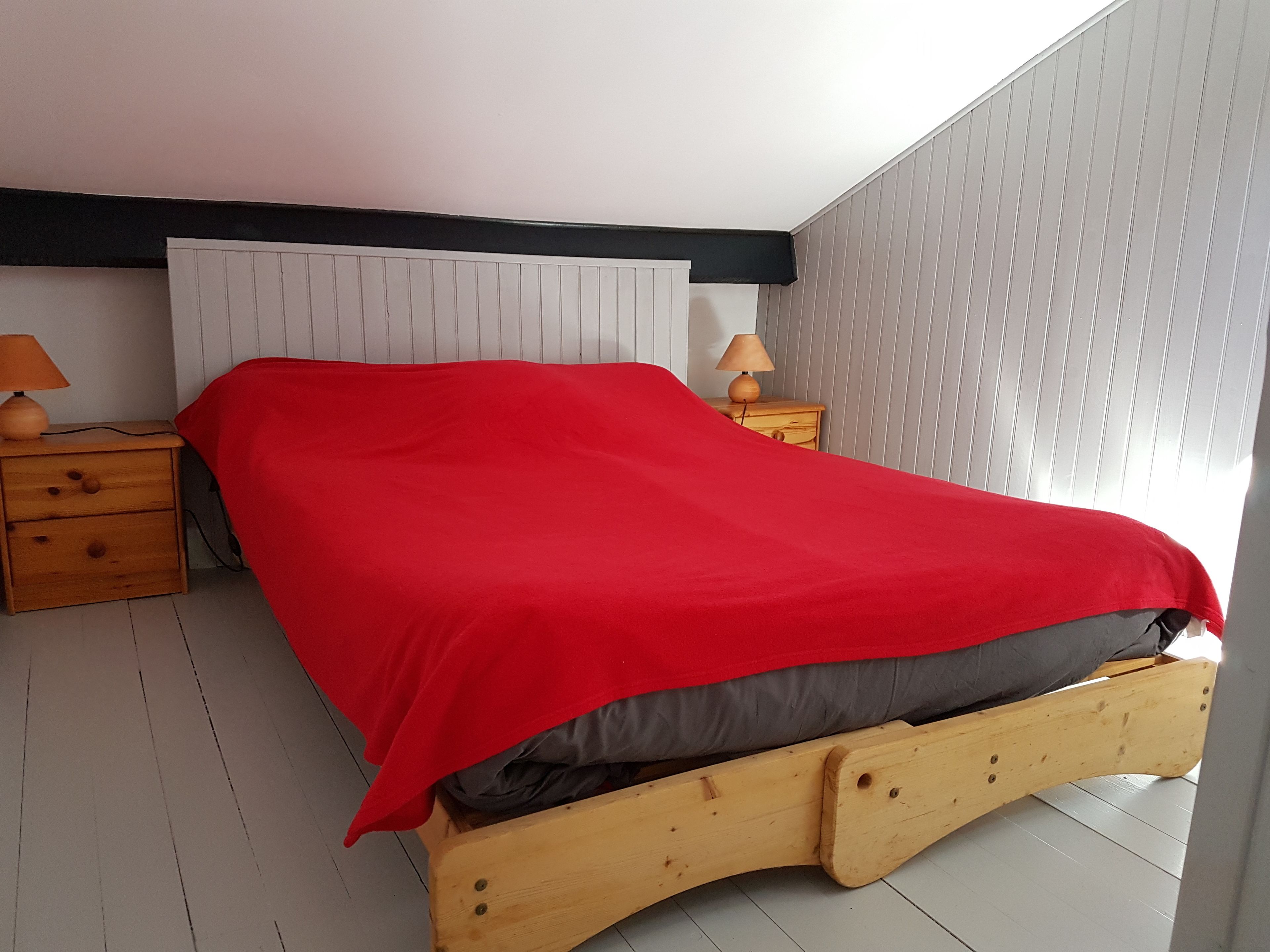 Une chambre à coucher avec un cadre de lit en bois, une tête de lit blanche et une couvre-lit rouge. On y trouve deux tables de chevet en bois, équipées de lampes et dotées d’un petit tiroir.