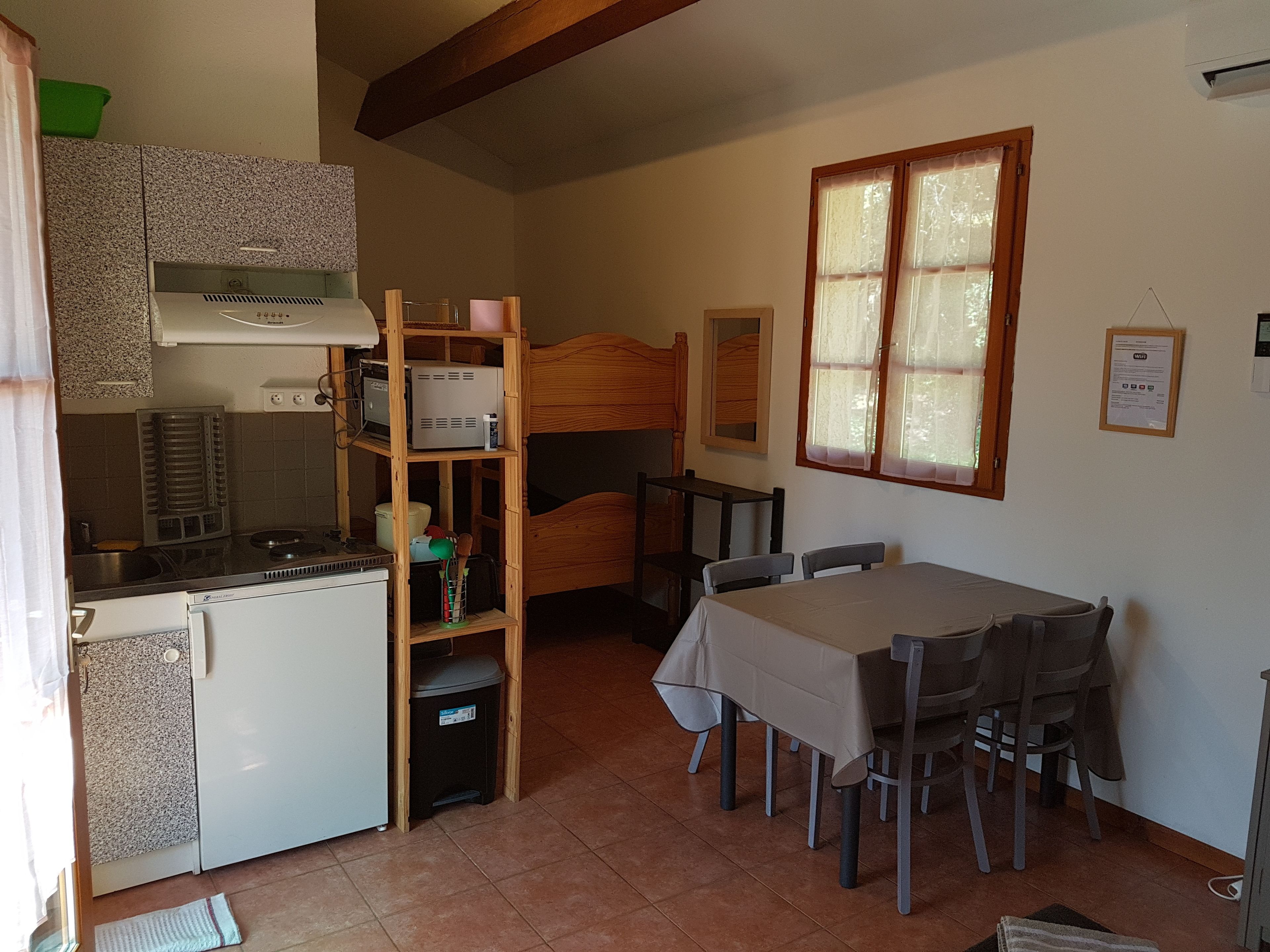 Una cocina compacta con estufa integrada, un refrigerador pequeño y una unidad de estanterías de madera. Hay una mesa de comedor con sillas y una ventana con cortinas.