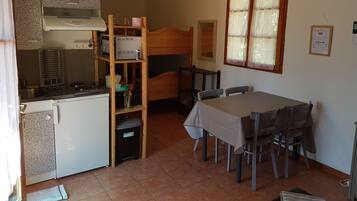 Una cocina compacta con estufa integrada, un refrigerador pequeño y una unidad de estanterías de madera. Hay una mesa de comedor con sillas y una ventana con cortinas.