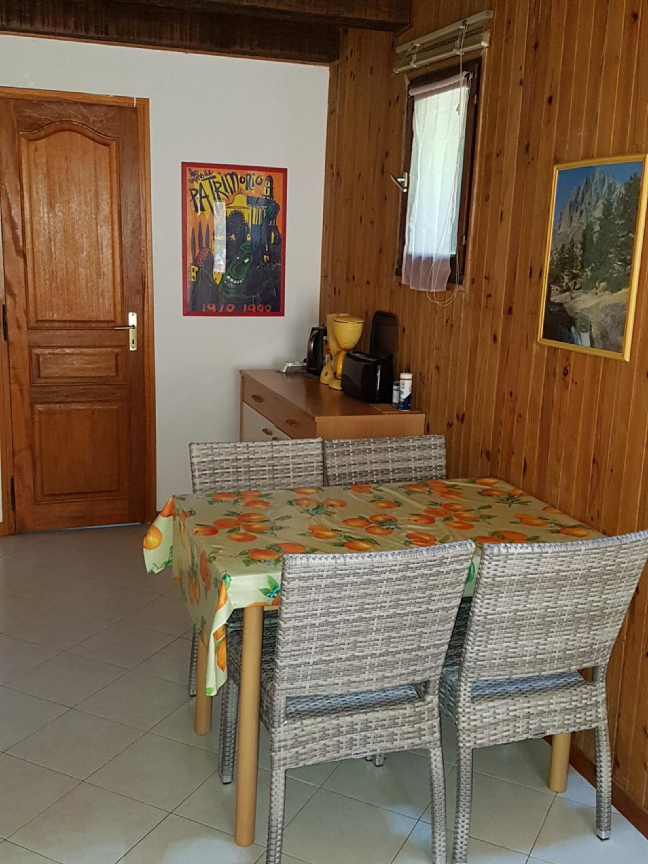 A dining area with a table covered by a fruit-patterned tablecloth, surrounded by wicker chairs.