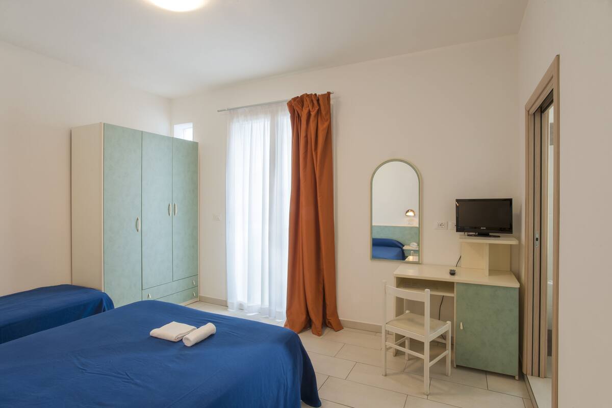 A bedroom with two beds, a wardrobe, a desk with a chair, a television, and a window with curtains.