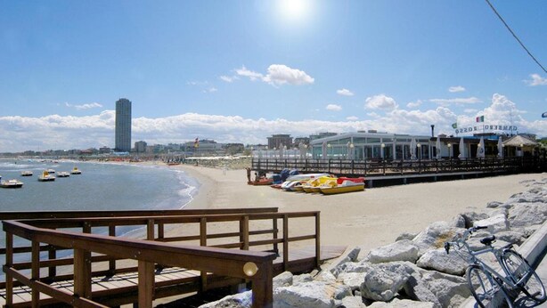 A beachside view with a wooden walkway, a restaurant with outdoor seating, and boats on the water. - Hotel Zamagna (Cesenatico)