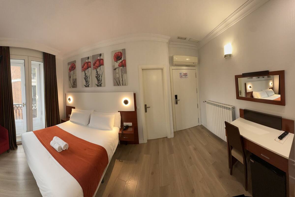 A hotel room with a bed, bedside table, nightstand, desk with chair, and a mirror.