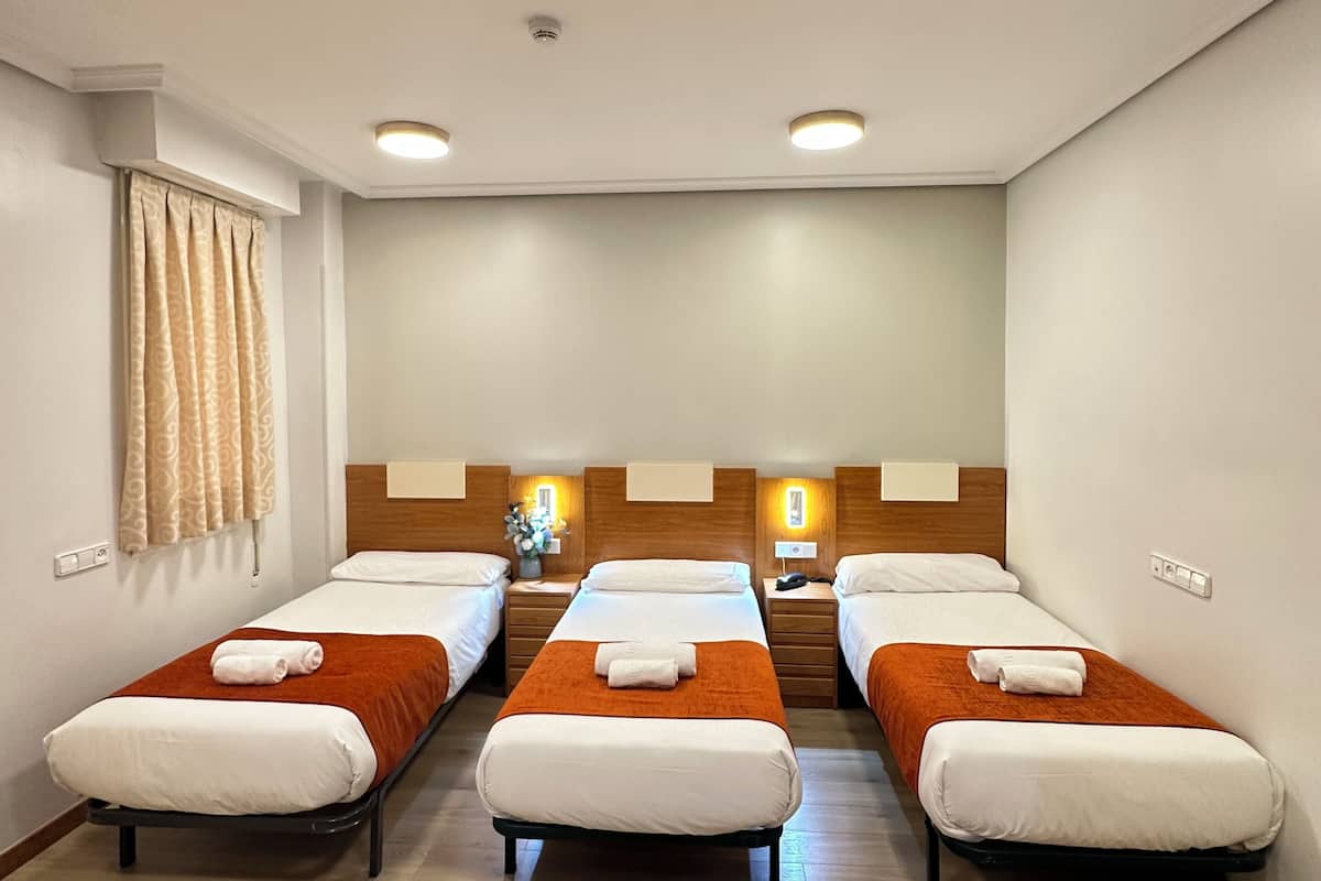 A hotel room with three single beds, a wooden headboard, a small table with a vase of flowers, and a mirror on the wall.