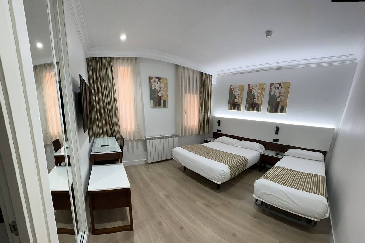 A hotel room with two single beds, a desk, a chair, a TV, and artwork on the wall.